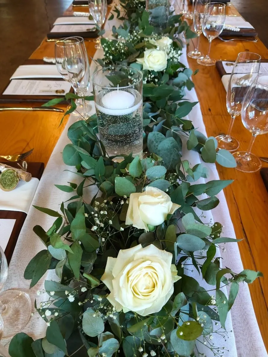 A garland of eucalyptus, white roses and baby's breath down a long timber table set with crystal glasses.
