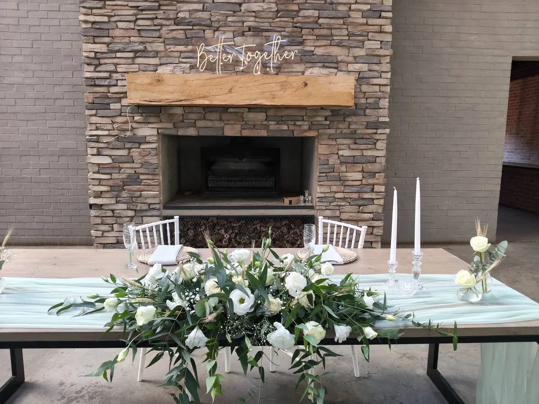 A bridal head table in front of a stone fireplace, with a low runner of white flowers and greenery and two white taper candles.