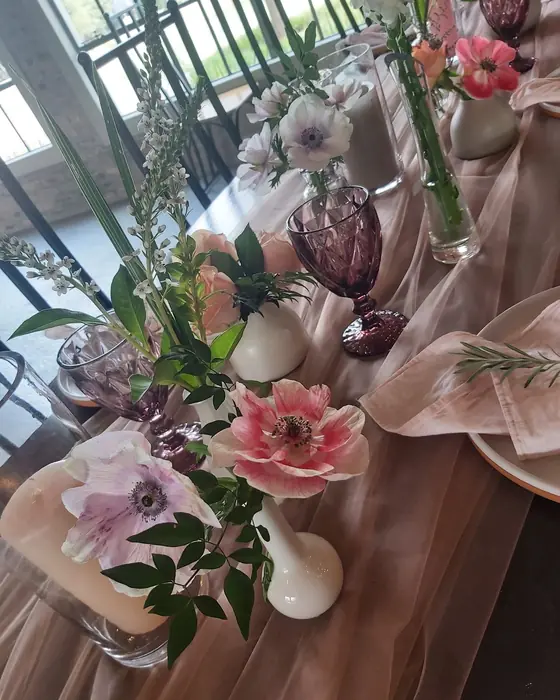Pale pink anemones and roses in a white vase on a blush tablecloth beside lilac glassware.