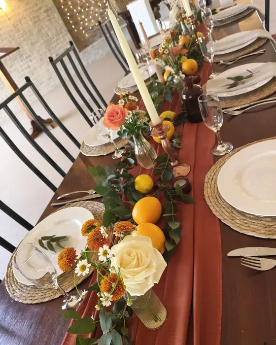 A rust runner with peach roses, oranges, lemons and taper candles between woven chargers.