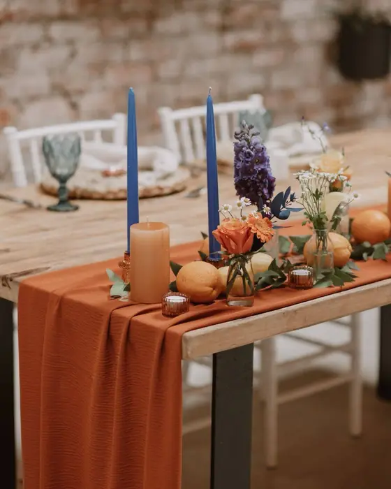 A terracotta tablecloth with blue taper candles, orange flowers and fruit in front of a brick wall.