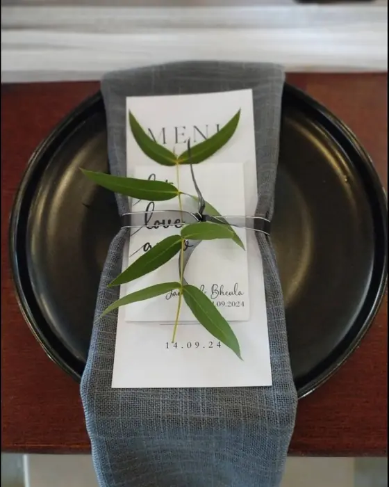 A menu card tied with an olive sprig on a grey napkin over a matte black charger.