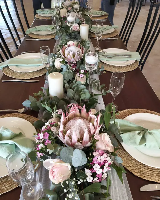 A long table with a runner of pink king proteas, roses and eucalyptus, and sage napkins on woven placemats.