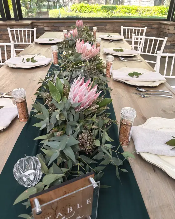 A forest green runner topped with proteas, eucalyptus and candles in glass holders, with a timber table number.