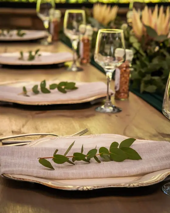 Place settings with eucalyptus sprigs on linen napkins and gold-rimmed chargers, with proteas behind.