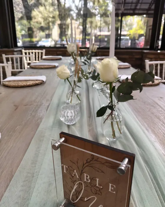 A white rose and greenery in a bud vase on a sage runner, with a timber table number reading Table One.
